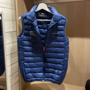 Blå dunväst från Swedemount  - Snygg blå dunväst från Swedemount med Nautic XPRNC RS65-tryck på bröstet. Västen har quiltad design, hög krage och dragkedja med röd detalj framtill. Perfekt för lager-på-lager och outdoor-häng. Logga även på ryggen för extra stilpoäng.