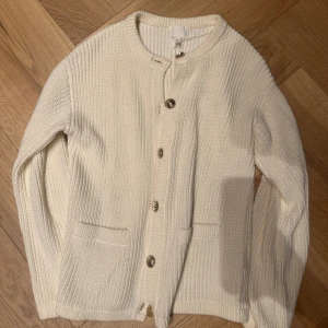 Vit stickad kofta med knappar - Säljer en stilren vit stickad kofta från H&M med rund hals och snygga silverfärgade knappar framtill. Koftan har två praktiska fickor och ribbad struktur som ger en cool och modern vibe. Perfekt att slänga på sig när det är lite kyligt ute.
