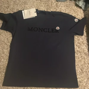 Mörkblå Moncler t-shirt med logga - Snygg mörkblå t-shirt från Moncler i storlek S. T-shirten har korta ärmar och ett stilrent Moncler-tryck på bröstet samt två broderade logopatchar på ärmen och bröstet. Materialet känns mjukt och skönt, perfekt för dig som gillar exklusiva märken.