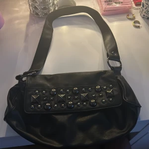 Svart axelväska med nitar - En svart axelväska i syntet med bred rem och lock dekorerat med silvriga nitar i olika former. Väskan har magnetlås och ett innerfack med dragkedja. Perfekt för dig som gillar en edgy stil och vill ha plats för det viktigaste. Jag använder inte den här väskan utan vill gärna bli av med den!