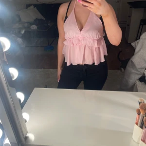 Ljusrosa volang topp med knyt - Supersöt ljusrosa topp med volanger och v-ringning. Toppen är ärmlös och har knyt i nacken för en snygg halterneck-look. Perfekt för dig som gillar en feminin och luftig stil. Passar till både jeans och kjol.