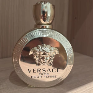 Versace Eros Pour Femme parfym - Lyxig parfymflaska i guld med Versaces ikoniska Medusa-huvud och snygga grekiska mönster runt kanten. Rund form med glänsande finish och guldfärgat lock. Parfymen är för kvinnor och flaskan känns riktigt exklusiv och elegant.