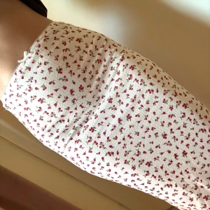 Vit blommig kjol från Pull&Bear - Supersöt vit kjol med små röda blommor från Pull&Bear. Kjolen är figurnära och har en liten rosett i midjan samt spetskant upptill. Perfekt för en romantisk look och passar extra bra till vår och sommar.