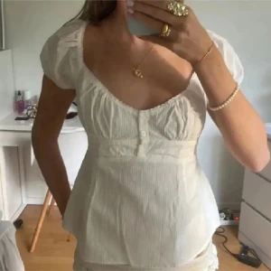 Vit blus med puffärm och knappar - Från Brandy Melville!Supersöt vit blus med korta puffärmar och v-ringad urringning. Toppen har små knappar framtill och lätt rynkade detaljer vid bysten. Figurnära passform och lätt, tunn bomullskänsla som passar perfekt till jeans eller kjol.