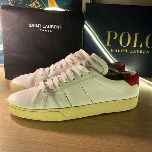 Saint Laurent skor  - Tha nu säljer jag mina snygga vita sneakers från Saint Laurent med röd detalj på hälen. Skorna är i skinn och har en beige sula med retrovibbar. Perfekta för dig som gillar stilrena och exklusiva sneakers med en twist. Skorna är i helt okej med sina skavanker men skickat får ni avgöra själva. Jag tar jättegärna byten och prisförslag. Med vänliga hälsningar Oscar