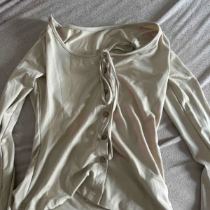 Beige långärmad topp med knyt - Säljer en beige långärmad topp med snygg knytning och knappar framtill. Modellen är offshoulder och har en slim passform som sitter tajt mot kroppen. Perfekt att styla med jeans för en chill och trendig look.
