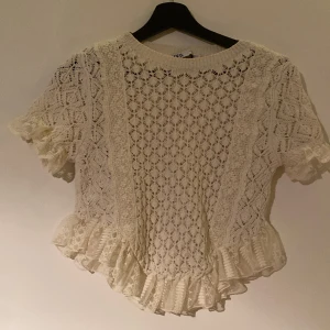 Vit spetsblus med volang från Zara - Superfin vit blus från Zara i storlek S, med detaljerad spets och stickat mönster. Blusen har korta ärmar, rund hals och volangkant längst ner som ger en romantisk vibe. Perfekt för dig som gillar boho och vill ha något extra till din outfit.