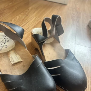 Svarta träskor med rem från Lola - Svarta träskor från Lola med klassisk design, tillverkade i skinn med träbotten. Skorna har en bred rem över foten och justerbar hälrem med spänne. Snygga detaljer med utskurna snitt framtill och rund tå. Lite skavanker eller vad man ska säga!