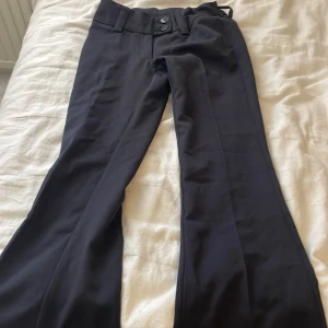 Svarta bootcut kostymbyxor från RYVLS - Snygga svarta kostymbyxor från RYVLS med bootcut-snitt och breda hällor för bälte. Byxorna har klassisk design med knapp och dragkedja framtill. Perfekta för dig som vill ha en stilren och trendig look.