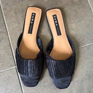 Svarta plisserade sandaler från Zara - Stilrena svarta sandaler från Zara med plisserad ovandel och fyrkantig tå. Snygg slip-in modell med platt sula, perfekt för en trendig look. Materialet har en lätt glansig finish som ger extra edge.helt nya 