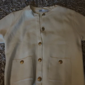 Beige kofta med guldknappar från Zara - Snygg beige kofta från Zara med raka linjer och dekorativa guldknappar framtill. Koftan har två stora fickor och en klassisk rund halsringning. Perfekt för lager-på-lager och enkel att styla till olika looks.