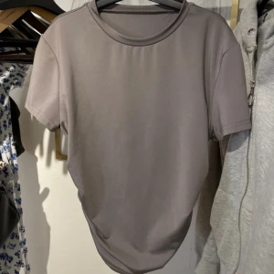 Grå croppad t-shirt  - Snygg grå t-shirt med korta ärmar och croppad passform. T-shirten har cool snörning på sidan som ger en unik look. Materialet känns mjukt och stretchigt, perfekt för dig som gillar stilrena plagg med en twist.