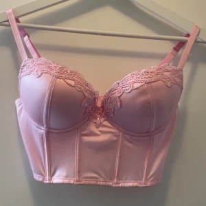 Rosa korsett-topp med spetsdetaljer - Supersöt ljusrosa korsett-topp med vadderade kupor och vacker spets längs bysten med glitter stenar. Justerbara axelband och hakknäppning bak. Storlek S. 