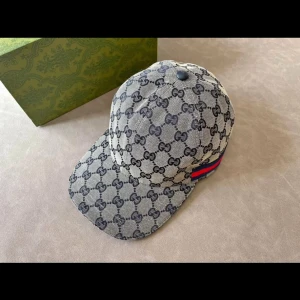 Gucci keps med GG-mönster - Snygg keps från Gucci med klassiskt GG-mönster i grått och svart. Kepsen har en böjd skärm och en röd-blå rand på sidan som ger en exklusiv touch. Perfekt accessoar för att lyfta din outfit.