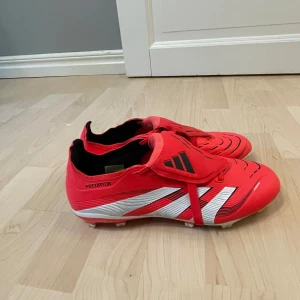 Adidas Predator röda fotbollsskor i storlek 39-40 utan lådan - Snygga Adidas Predator fotbollsskor i rött med vita och svarta detaljer. Skorna har klassisk snörning, täckt plös och mönstrad ovandel. Utrustade med fasta dobbar för gräsplan. Perfekta för dig som vill sticka ut på planen.