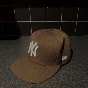 Brun New Era NY keps - Snygg brun New Era keps med klassiskt New York Yankees-logo broderat i vitt framtill. Kepsen har rak skärm och broderad New Era-logga på sidan samt MLB-logga bak. Tillverkad i ett tåligt tygmaterial, perfekt för en streetwear-look.