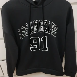 Svart hoodie Los Angeles 91 från SHEIN - Svart hoodie från SHEIN med trycket 'Los Angeles 91' i vitt på framsidan. Tröjan har huva med snörning och långa ärmar. Mjuk insida och normal passform, perfekt för chill dagar. Materialet är troligtvis polyester. Storlek S (EU 36). Tyget har lite defekter pga tvätt, kolla sista bilden.