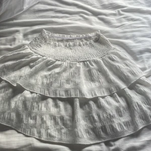 Vit volangkjol med smock - Supersöt vit kortkjol med dubbla volanger och smockad midja. Kjolen är gjord i ett lätt, krinklat bomullstyg som ger en chill och luftig känsla. Perfekt för varma dagar och har en riktigt fräsch vibe med sin ljusa ton och volangdetaljer.