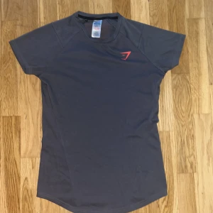 Mörkgrå Gymshark t-shirt med röd logga - Sportig mörkgrå t-shirt från Gymshark i storlek S. T-shirten har korta ärmar, rund hals och en röd Gymshark-logga på bröstet samt ett stort rött tryck längs ryggen. Materialet är en mix av bomull och återvunnen polyester med lite stretch för skön passform. Tröjan är i mycket bra skick.