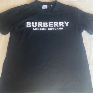 Svart Burberry t-shirt med tryck - Svart t-shirt från Burberry med stort vitt tryck på bröstet där det står 'BURBERRY LONDON ENGLAND'. Klassisk rund hals och korta ärmar. Tillverkad i mjuk bomull som är skön mot huden. Perfekt för dig som gillar stilrena och ikoniska plagg.