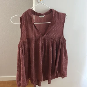 Rostbrun ärmlös blus från Only - Säljer en luftig rostbrun blus från Only med v-ringning och volangdetaljer. Blusen är ärmlös och har ett diskret prickigt mönster i tyget samt transparenta partier på ryggen. Perfekt för dig som gillar en bohemisk och avslappnad stil.