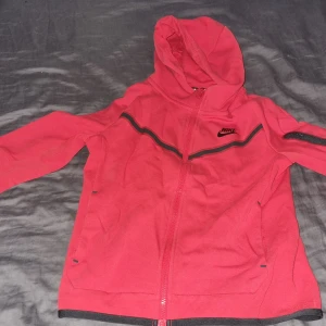 Röd Nike tech hoodiejacka med dragkedja - Nike hoodiejacka i röd med svarta detaljer och hel dragkedja. Jackan har huva, långa ärmar och två fickor framtill. Tillverkad i mjukt bomullsmaterial, perfekt för chill dagar eller när du vill ha en sportig look. Pris kan diskuteras skriv vid minsta fundering😊