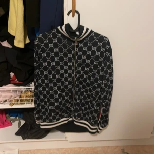 Zip hoodie från Gucci, Small - Gucci bomberjacka i svart med vit GG-monogram över hela jackan. Jackan har dragkedja framtill, ribbade muddar och krage med vita ränder. Tillverkad i bomull och polyester för en skön och sportig känsla. Perfekt för dig som vill sticka ut med lyxig streetstyle.