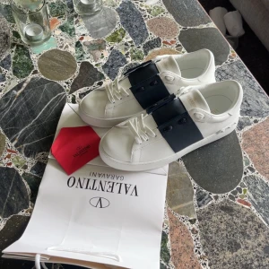 Valentino Garavani Open sneakers vit/blå Storlek 43 - Snygga Valentino Garavani Open sneakers i vitt skinn med bred marinblå rem över vristen och ikoniska nitar baktill. Klassisk rund tå, platt sula och vita snören. Lyxig och stilren design som sticker ut. Kommer utan Skokartong