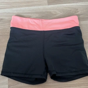 Svarta sportshorts med rosa midja H&M - Sportiga svarta shorts från H&M med bred, ljusrosa hög midja. Stretchigt syntetmaterial som sitter tight och är perfekt för träning. Snygg kontrast mellan svart och rosa, med diskret H&M Sport-logga på insidan. Passar dig som gillar att röra på dig.