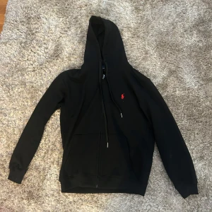 Svart hoodie från Polo Ralph Lauren - Svart hoodie från Polo Ralph Lauren med röd broderad logga på bröstet. Tröjan har huva med snörning, dragkedja framtill och känguruficka. Tillverkad i mjuk bomull, perfekt för en chill och stilren look.