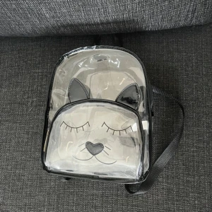 Transparent ryggsäck med kattmotiv - Säljer en liten transparent ryggsäck med svart kant och justerbara axelband. Framsidan har ett gulligt kattansikte med stängda ögon och hjärtformad nos, samt svarta öron i syntetmaterial. Perfekt för dig som gillar unika accessoarer med djurtema.