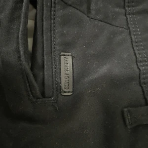 Svarta chinos från Jack & Jones - Svarta chinos från Jack & Jones i klassisk modell med dragkedja och knapp framtill. Byxorna har snedställda fickor och är tillverkade i bomull med en skön passform. Perfekta för en clean och stilren look.