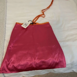 Rosa satin kjol med orange kant - Snygg rosa kortkjol i satin med en cool orange kant upptill och matchande orange knytband. Kjolen har en slät yta och en enkel, rak passform som känns både trendig och lekfull.