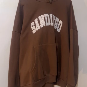 Brun hoodie med SAN DIEGO-tryck - Säljer en brun hoodie med vit SAN DIEGO-text på bröstet. Tröjan har en stor magficka, huva och långa ärmar. Perfekt för chill dagar och har en loose passform. Materialet känns som mjuk bomull och polyester, riktigt skön att bära.