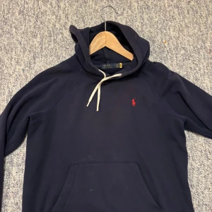 Mörkblå hoodie från Polo Ralph Lauren - Snygg mörkblå hoodie från Polo Ralph Lauren med klassisk röd logga på bröstet. Tröjan har huva med vit snörning och en stor magficka framtill. Tillverkad i mjuk bomull, perfekt för chill dagar eller när du vill ha en clean streetstyle-look.