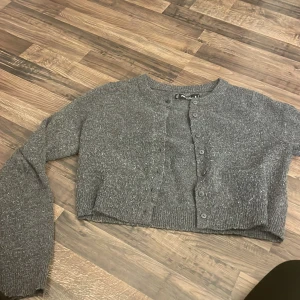 Grå croppad kofta från FB Sister - Grå croppad kofta från FB Sister med rund hals och långa ärmar. Tröjan är stickad och har knappar framtill. Perfekt för lager-på-lager och chill stil. Materialet är mjukt och ger en mysig känsla. Passar dig som gillar enkel och clean look.