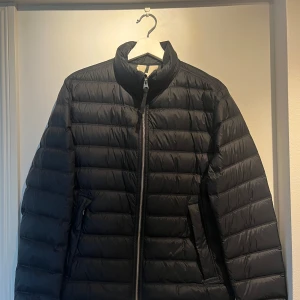 Svart pufferjacka med dragkedja - Snygg svart pufferjacka med horisontella sömmar och hög krage. Jackan har hel dragkedja framtill och två fickor på sidorna. Perfekt för höst. Storleken är 48 men känns som 46  