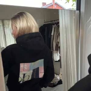 Svart hoodie från Calvin Klein - Svart hoodie från Calvin Klein med stor färgglad logopatch på ryggen. Tröjan har huva med snörning och en klassisk magficka framtill. Mjuk och skön bomullskänsla, perfekt för chill dagar. Passformen är loose och ärmarna är långärmade.