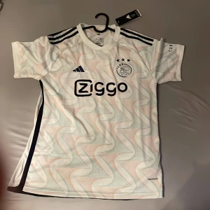 Ajax vit kortärmad fotbollströja Adidas - Ajax fotbollströja från Adidas i vit med subtilt zigzag-mönster i ljusa toner. Tröjan har svarta detaljer, tre ränder på axlarna och Ziggo-logga på bröstet. Ajax-emblem med tre stjärnor på vänster sida. Tillverkad i lätt och ventilerande material.
