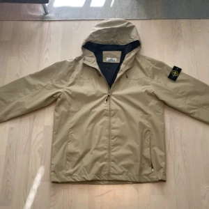 Beige vindjacka från Stone Island - Snygg beige vindjacka från Stone Island med huva och dragkedja framtill. Jackan har klassisk patch på ärmen och två fickor med dragkedja. Perfekt för dig som vill ha en stilren och funktionell jacka med streetkänsla.