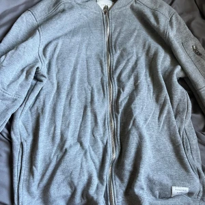 Grå zip hoodie från Jack & Jones - Säljer en stilren grå hoodie från Jack & Jones Core i storlek L. Tröjan har dragkedja framtill, huva och långa ärmar. Mjuk bomullskänsla och enkel design med diskret logotyp på sidan. Perfekt för chill dagar eller när du vill ha en clean look.