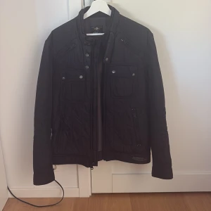 Svart quiltad jacka med dragkedja - Svart quiltad jacka från replay med coola detaljer som tryckknappar, dragkedjor och flera fickor framtill. Jackan har hög krage och lång ärm, perfekt för dig som gillar en stilren och sportig look.