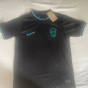 Svart Brasilien fotbollströja Nike - Snygg svart Brasilien fotbollströja från Nike med blå detaljer på krage och ärmslut. Tröjan har korta ärmar och är tillverkad i ett lätt, sportigt material. Brasilien-emblemet och Nike-loggan syns framtill i blått. Perfekt för dig som älskar fotboll och vill sticka ut.