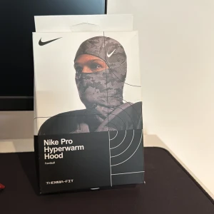 shiesty Pro Hyperwarm Hood  - Nike Pro Hyperwarm Hood är en svart, tight huva med camouflagemönster, designad för att bäras under hjälm eller ensam. Tillverkad i polyester och elastan med Therma-FIT-teknologi som håller dig varm och bekväm. Perfekt för fotboll och träning under kyliga dagar.