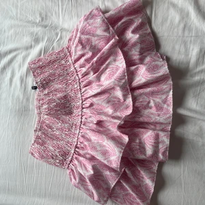 Rosa volangkjol från EET x TIGER - Supersöt kortkjol från MEET x TIGER i storlek S i ljusrosa och vitt med mönstrad design. Kjolen har bred smock i midjan med volanger. Den har även inbyggda shorts. Kommer inte till användning, köpt för 2 år sen och är i mycket bra skick då den inte använts mycket alls. 