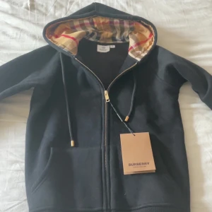 Svart Burberry hoodie med rutig huva - Svart hoodie från Burberry med klassisk rutig insida i huvan i beige, rött och grönt. Tröjan har dragkedja framtill, snörning vid huvan och två fickor. Snygga guldfärgade detaljer på snörningen och lyxig känsla. Perfekt för dig som gillar stilrena märkesplagg.