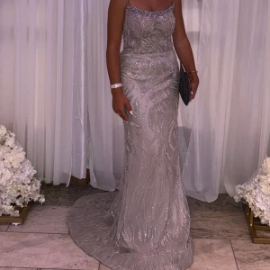 Glittrig långklänning i silver - Säljer en magisk långklänning i silver med glittriga detaljer och mönster. Klänningen har en figurnära passform, smala axelband och en elegant släp. Materialet är skimrande och ger en lyxig känsla, perfekt för bal eller fest.
