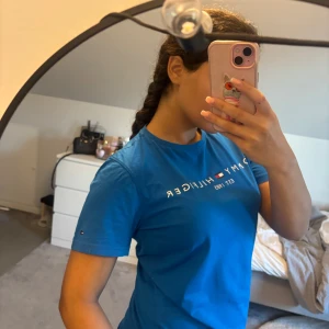 Blå Tommy Hilfiger t-shirt i bomull - Snygg blå t-shirt från Tommy Hilfiger med klassisk logga och text 'EST. 1985' framtill. Tillverkad i mjuk ekologisk bomull, rund hals och korta ärmar. Liten Hilfiger-flagga på ärmen och cool detalj med röd-vit-blå band i nacken.