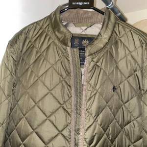 Olivgrön quiltad jacka från Morris - Snygg olivgrön quiltad jacka från Morris med klassisk bomberkrage och diskret broderad logga på bröstet. Jackan har dragkedja framtill och ett stilrent rutmönster. Perfekt för dig som gillar en clean och tidlös look.