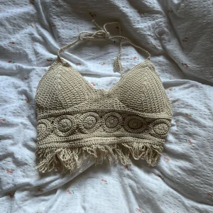 Beige virkad croppad linnetopp - Säljer en beige virkad topp med knyt i nacken och snygga virkade detaljer. Toppen har fransar längst ner och vadderade kupor på insidan. Perfekt för sommarens festivaler eller strandhäng!